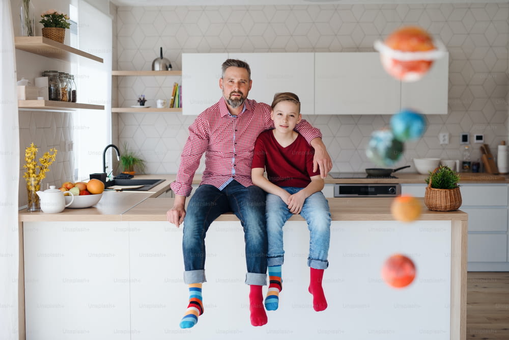 Mature father and small son with colorful socks sitting on kitchen counter indoors, looking at camera.