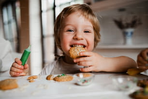 Um pequeno menino feliz sentado à mesa, decorando e comendo bolos em casa.