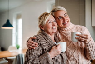 A happy senior woman and adult daughter with coffee at home, looking out of window.