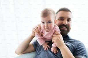 Portrait of young father holding baby daughter sitting indoors on a sofa.