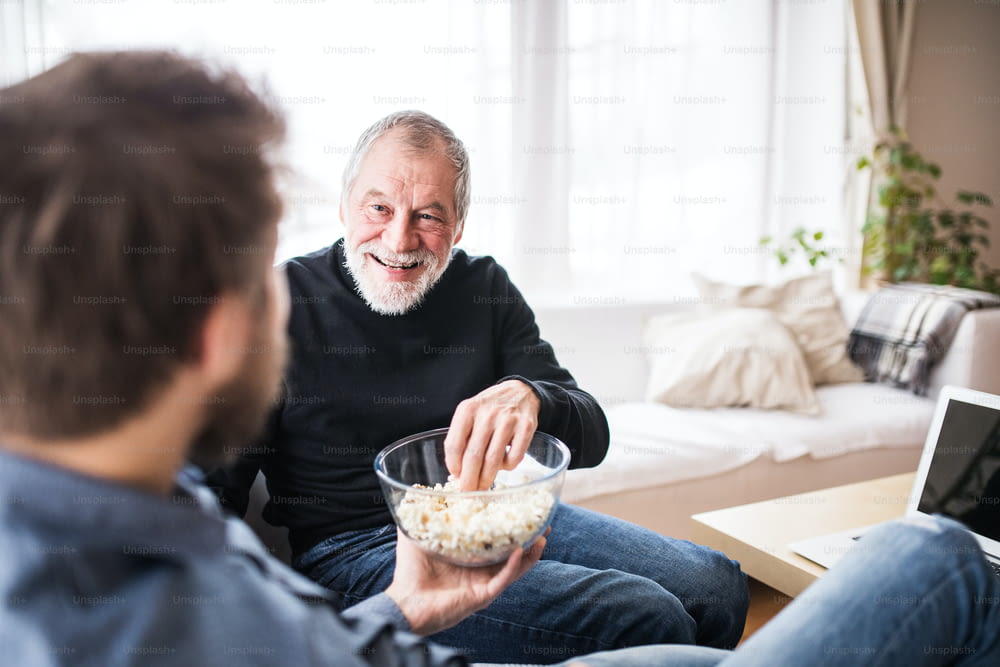 Hipster son and his senior father at home, eating popcorn. Two generations indoors.
