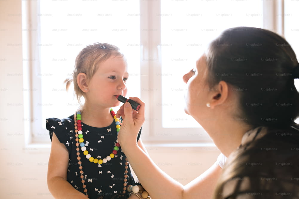 Beautiful young mother painting lips of her cute little daughter with lipstick.