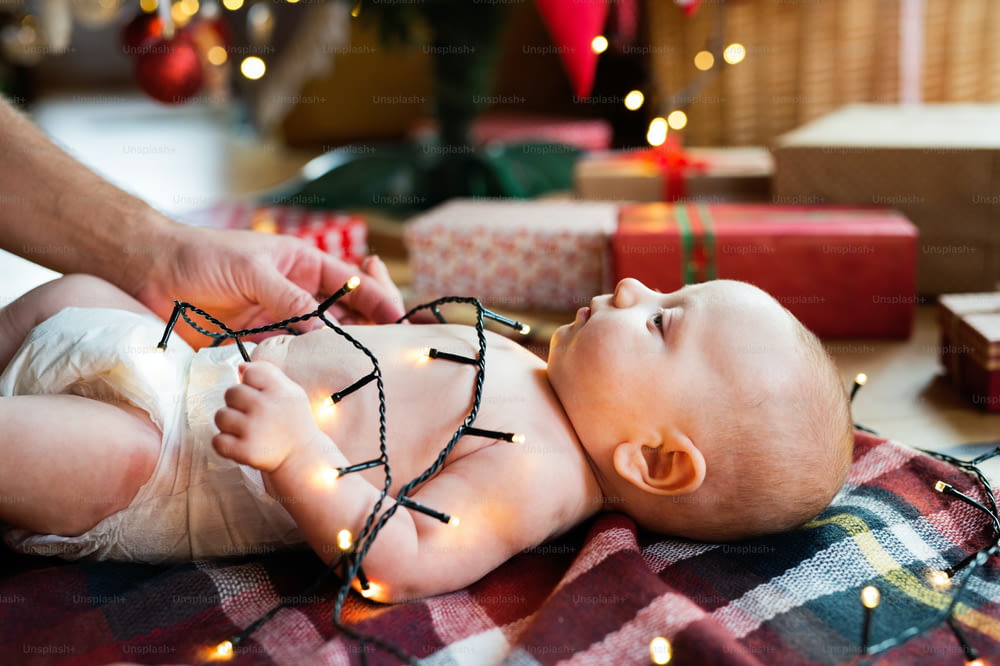 Unrecognizable father with baby boy at Christmas time. Baby lying on the blanket on the floor., tangled in the chain of lights.