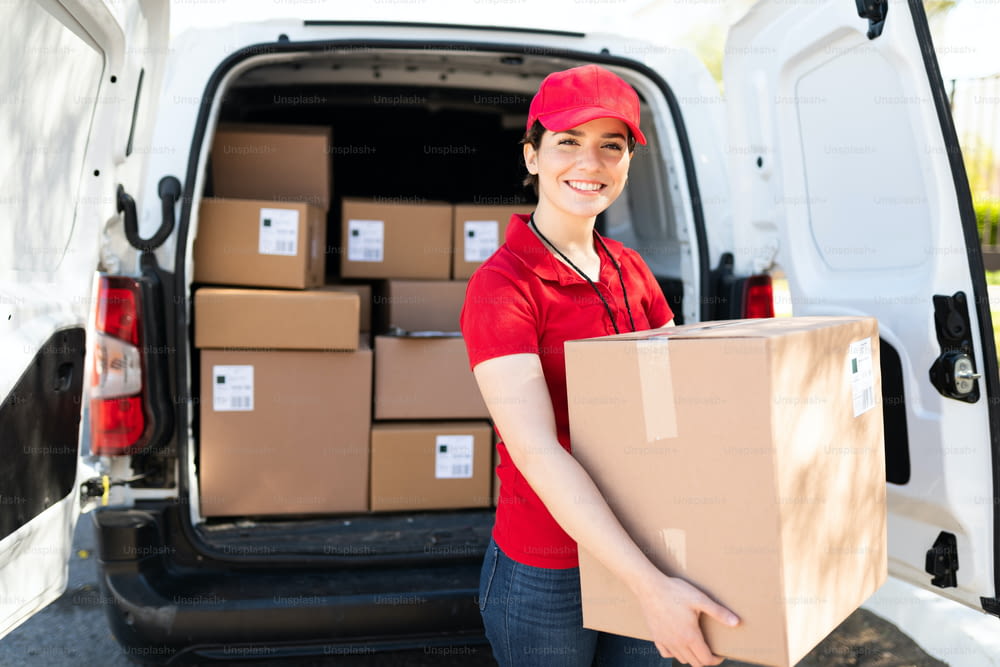 Portrait of an attractive caucasian female worker in a red uniform unloading packages while smiling and standing next to a delivery van with a lot of boxes and parcels