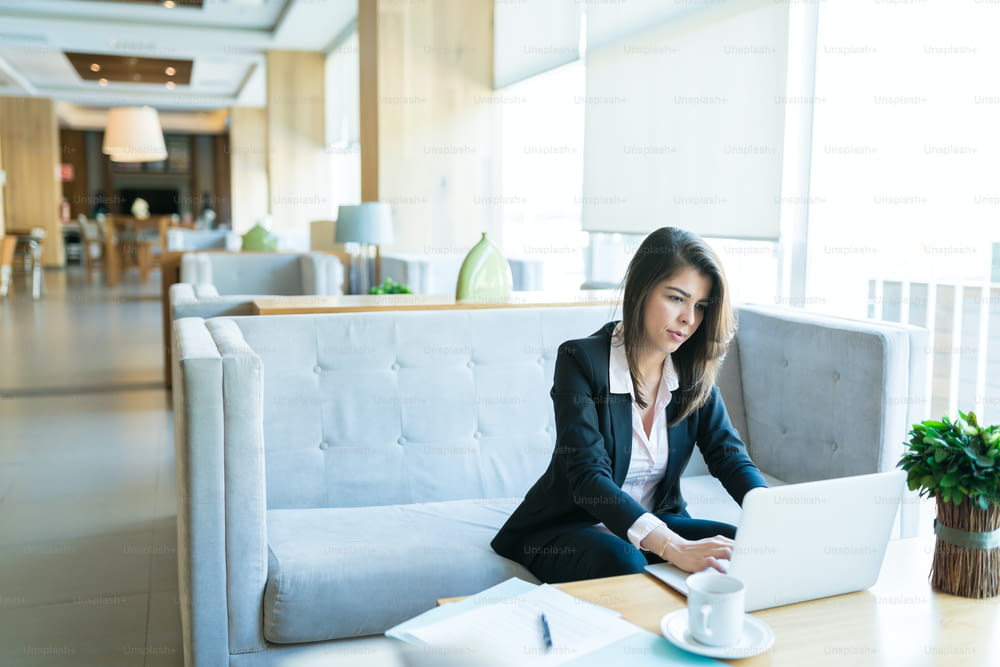 Confident Latin businesswoman using laptop while sitting with coffee and documents at table in hotel lobby