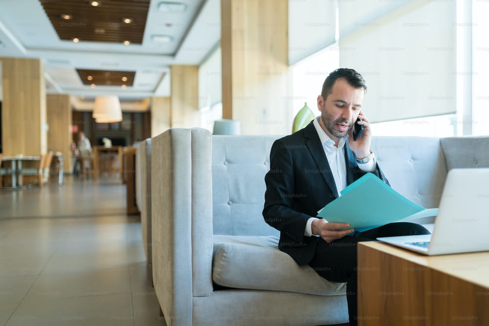 Handsome mid adult businessman busy on call while reading business plan from file in hotel lobby