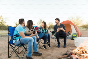 Des amis heureux avec des bouteilles d’alcool assis sur des chaises au camping