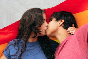 Um jovem casal de meninas se beijando com uma bandeira do orgulho gay em um parque em um dia ensolarado, celebrando o dia do orgulho e se apaixonando.