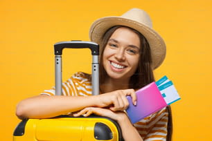 Concepto de viaje. Joven estudiante viajera feliz con pasaporte con billetes de avión y maleta, aislada sobre fondo amarillo