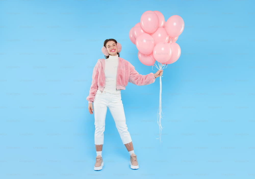 Young positive girl wearing pink fluffy winter bomber and ear warmers, standing with bunch of pink balloons, celebrating her birthday, isolated on blue background