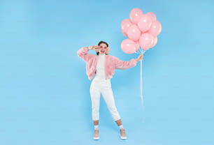 Retrato de cuerpo entero de una joven mujer feliz con bomber rosa esponjoso y calentadores de orejas, de pie con globos de cumpleaños rosas, aislados sobre fondo azul