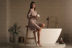Portrait of satisfied relaxed woman with cup of herbal tea in silk robe resting after body massage in spa and beauty salon sitting on wooden stool in luxury bathroom with white bathtub on background