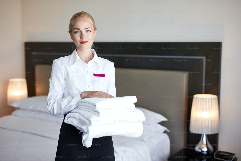 Attraktives Dienstmädchen mit Handtüchern in den Händen, charmante Frau steht im ordentlichen Zimmer, bereitet ein Zimmer für die Gäste vor