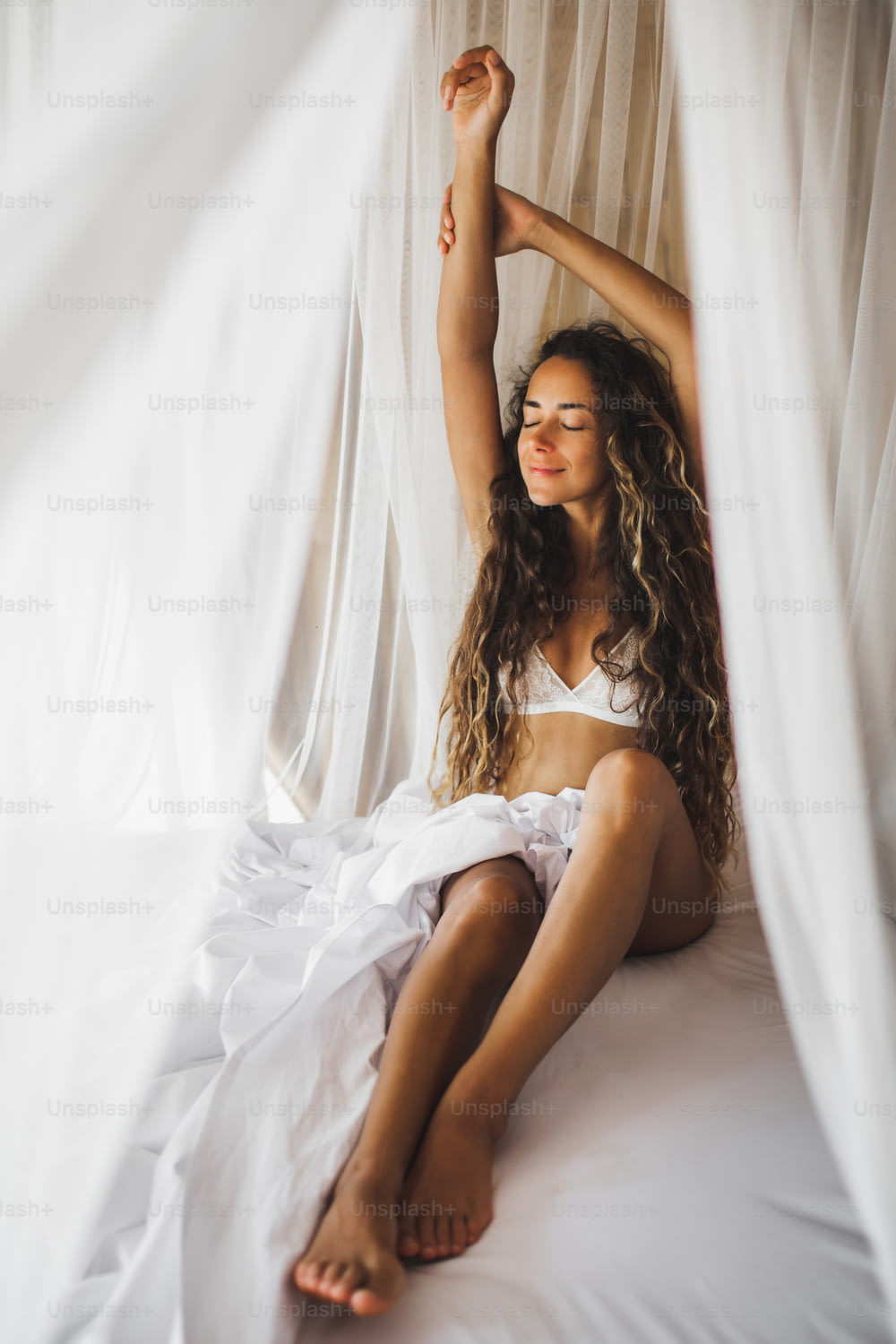 Beautiful tanned young woman awakening in white bed. Happy wake up and start new day. Leisure and rest. Wellbeing and carefree  concept. Long curly hair. Stay home.