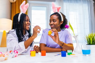 multiracial two women painting eggs with cute pink rabbit costume ears in apartment .
