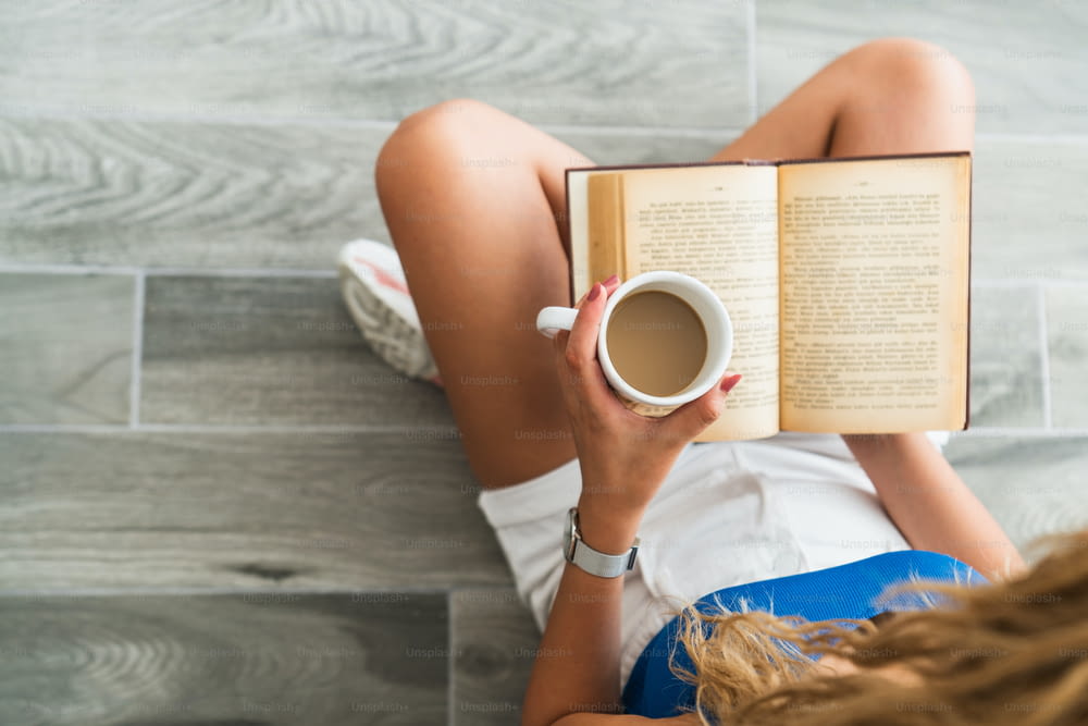 Ragazza intellettuale seduta sul pavimento e bere una tazza di caffè, mentre sta leggendo un libro. Foto di alta qualità