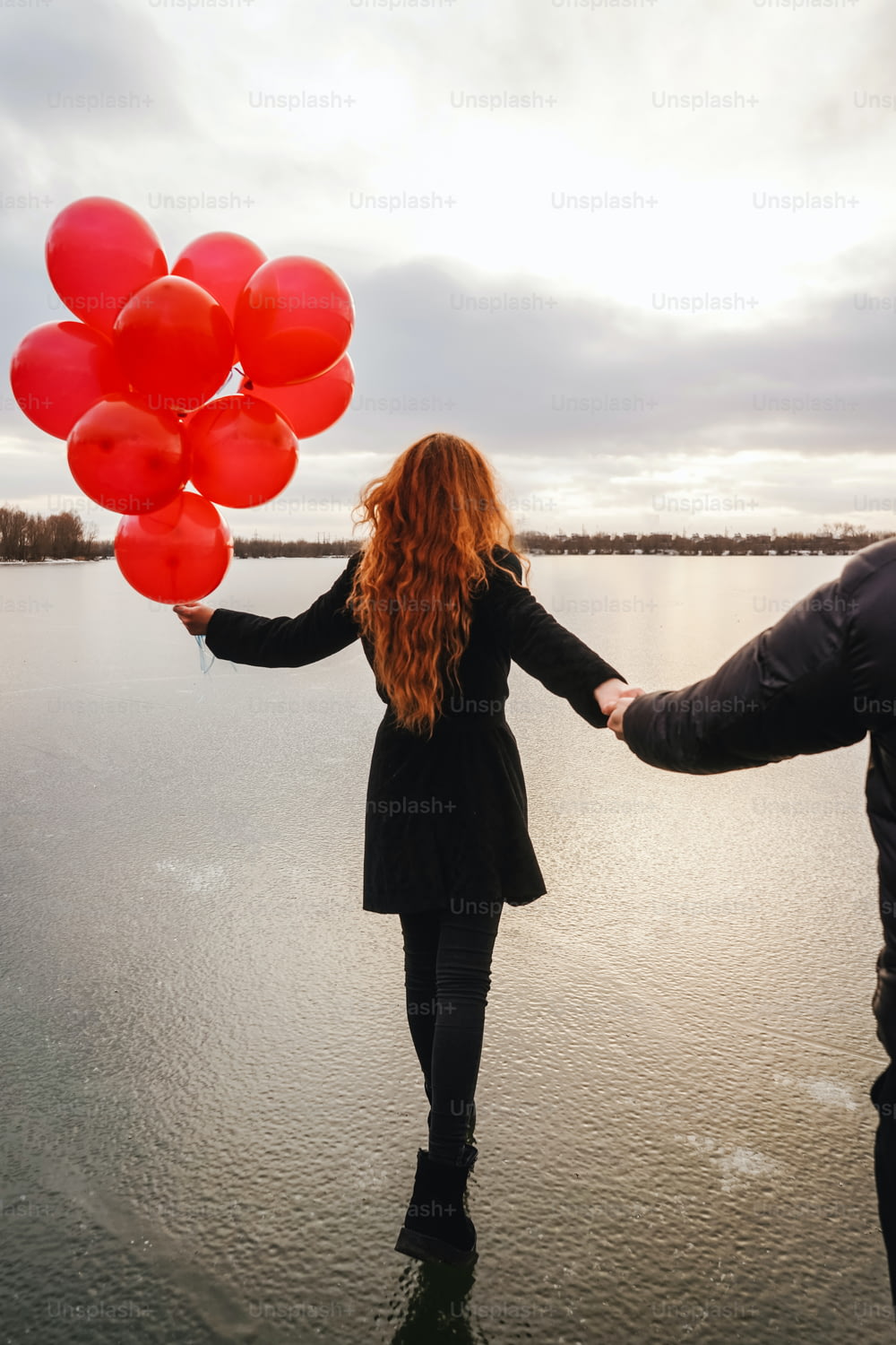 Love couple with red balloons outdoor in winter ice river. Sunset light, back view. Black casual clothes. Follow me concept, girl walks in front of the guy and holds the guy's hand.