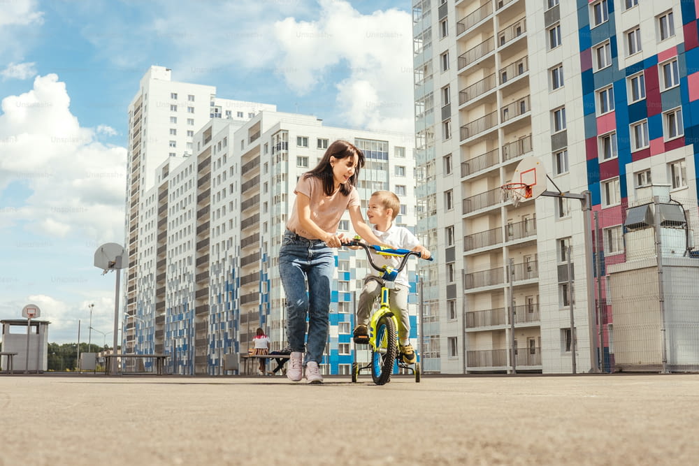 boy rides a bike with his mother. Houses on background, summer.