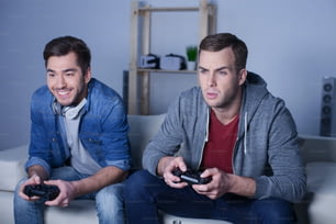 Attractive man is winning his friend in play-station. He is sitting on sofa and smiling. Another man is looking at monitor with seriousness