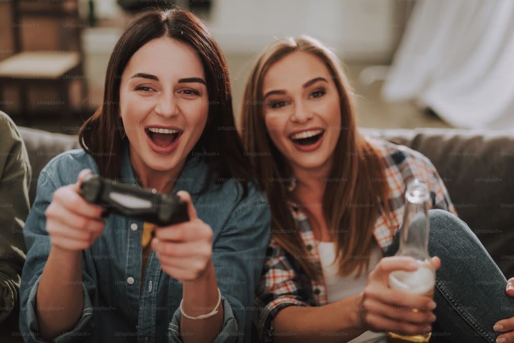 Portrait of excited young lady using joystick while her friend holding bottle of beer and laughing