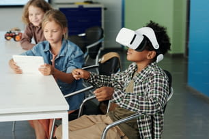 Vista laterale del simpatico scolaro afroamericano in auricolare vr che controlla il robot virtuale mentre il suo compagno di classe guarda l'azione sullo schermo del tablet