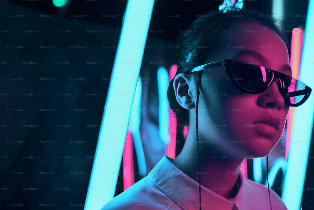 Retrato da jovem adolescente asiática em óculos de sol elegantes em forma de crescente, em luz neon vermelha anf azul. Conceito de retrato cibernético e futurista