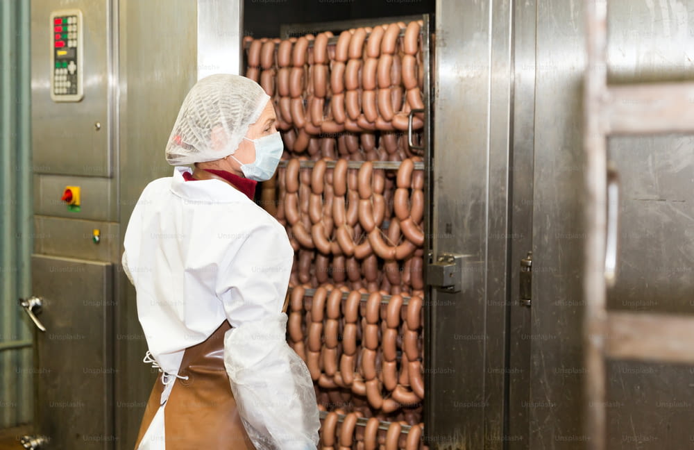 Woman checking quality of cooked smoked sausages in professional baking machine at food production factory