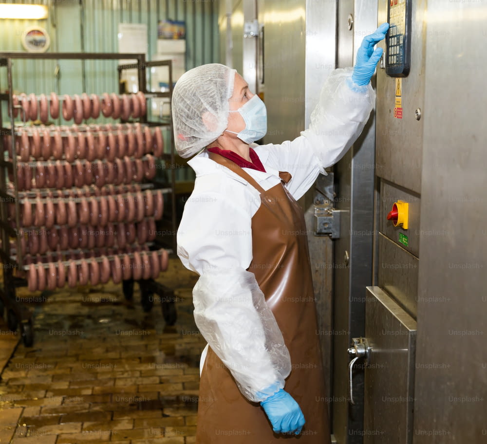 Competent workwoman programming sausage baking machine at food production factory