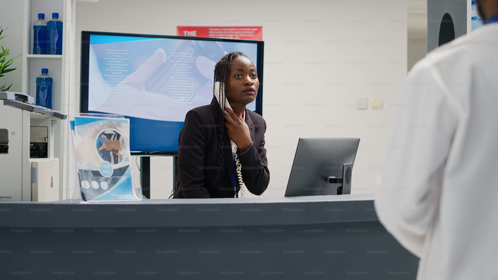 Mujer afroamericana contestando llamadas telefónicas fijas en el mostrador de recepción, trabajando en el hospital para brindar apoyo a los pacientes. Adulto joven uing teléfono con cable en la recepción del centro de salud.