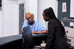 African american medical staff analyzing checkup report with patient insurance support on papers. Nurse and receptionist working on checkup visit appointments at hospital reception desk, healthcare.