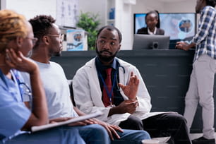 Hombre consultor del equipo médico afroamericano sentado en la sala de espera en el vestíbulo de recepción de las instalaciones. Médico y asistente hablando con el paciente sobre el diagnóstico y tratamiento de la enfermedad, apoyo de atención médica.