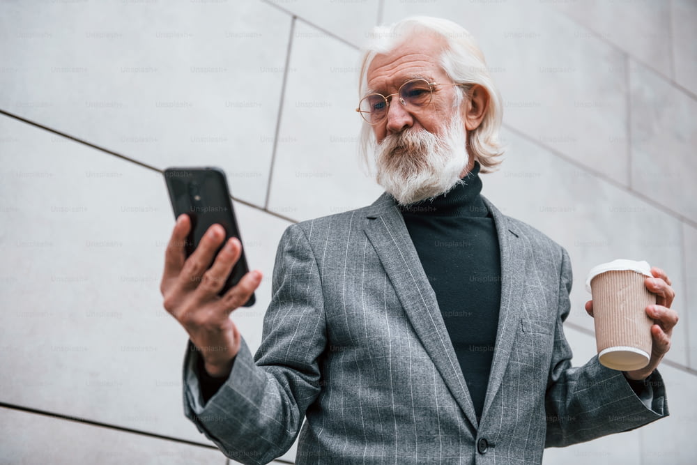 Usando smartphone. Empresário sênior com roupas formais, cabelos grisalhos e barba ao ar livre.