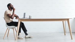 Vista lateral de um jovem negro pensativo usando laptop, sentado na mesa, pensando em algo, trabalhando on-line em home office, espaço livre. Homem afro-americano sonhador que tem trabalho remoto