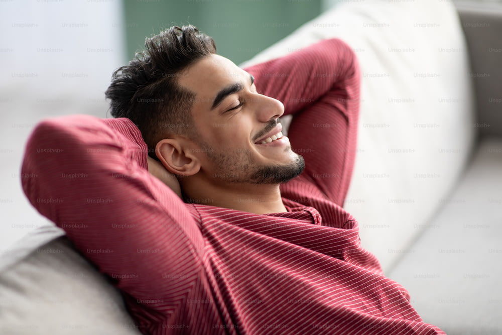 Closeup of peaceful muslim guy with closed eyes and hands behind his head reclining on couch at home, arab man enjoying weekend alone, dreaming about wealthy life, side view, copy space