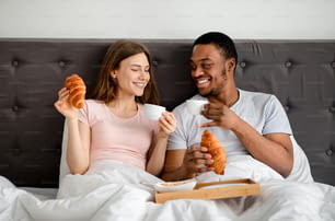 Millennial multiracial couple having breakfast in bed together, drinking coffee with croissants, spending honeymoon at hotel. Multiethnic newlyweds enjoying romantic morning with tasty meal