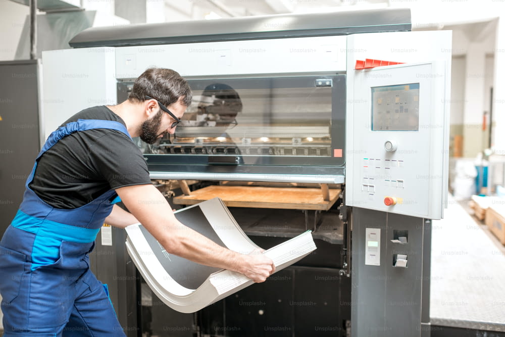 Worker in unifrom getting printed sheets from the offset machine at the printing manufacturing