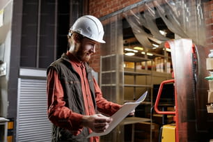 Side view portrait of bearded worker wearing hardhatat factory and looking at documents