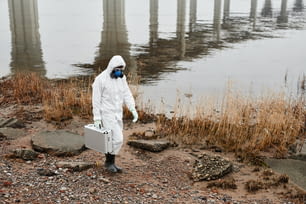 Full length portrait of worker wearing hazmat suit walking by water outdoors carrying samples case, industrial waste concept, copy space