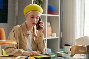 Junge Unternehmerin in stilvoller Freizeitkleidung, die auf dem Handy spricht und während der Arbeit im Studio die Tasten des Taschenrechners drückt