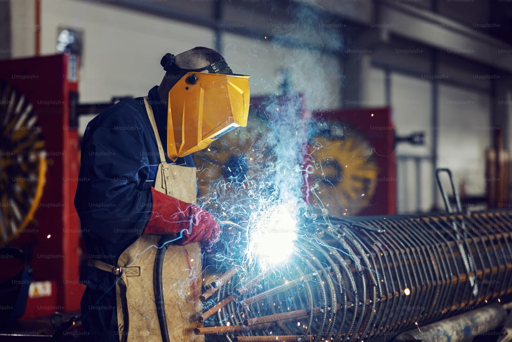 A heavy industry metal worker is welding metal framework with a welding machine in a factory.