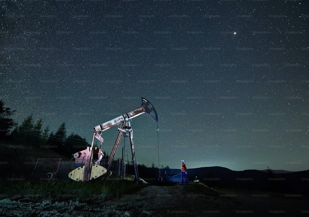Homem de petróleo de pé perto de jack bomba de petróleo sob o belo céu noturno com estrelas. Engenheiro de petróleo controlando o trabalho de balancim bomba de óleo à noite em campo de petróleo. Conceito de extração de petróleo.