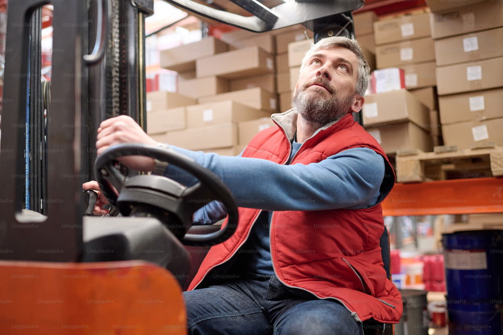 Mature warehouse worker looking on high shelves during loading cargo with forklift
