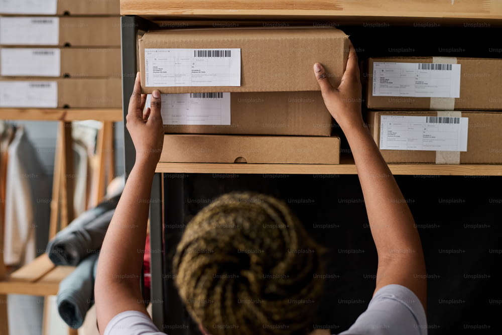 Arme einer jungen schwarzen Frau, die verpackte Pappkartons mit Waren sortiert, die von Online-Kunden bestellt wurden, und eine aus dem Holzregal nehmen