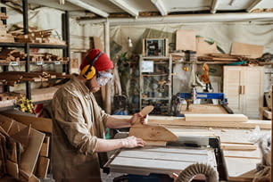 Young male carpenter wearing safety glasses and hearing protection headphones cutting wood plank with table saw