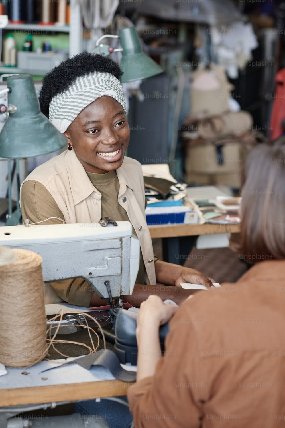 African young tailor smiling to her colleague while they talking during their sewing work in the workshop