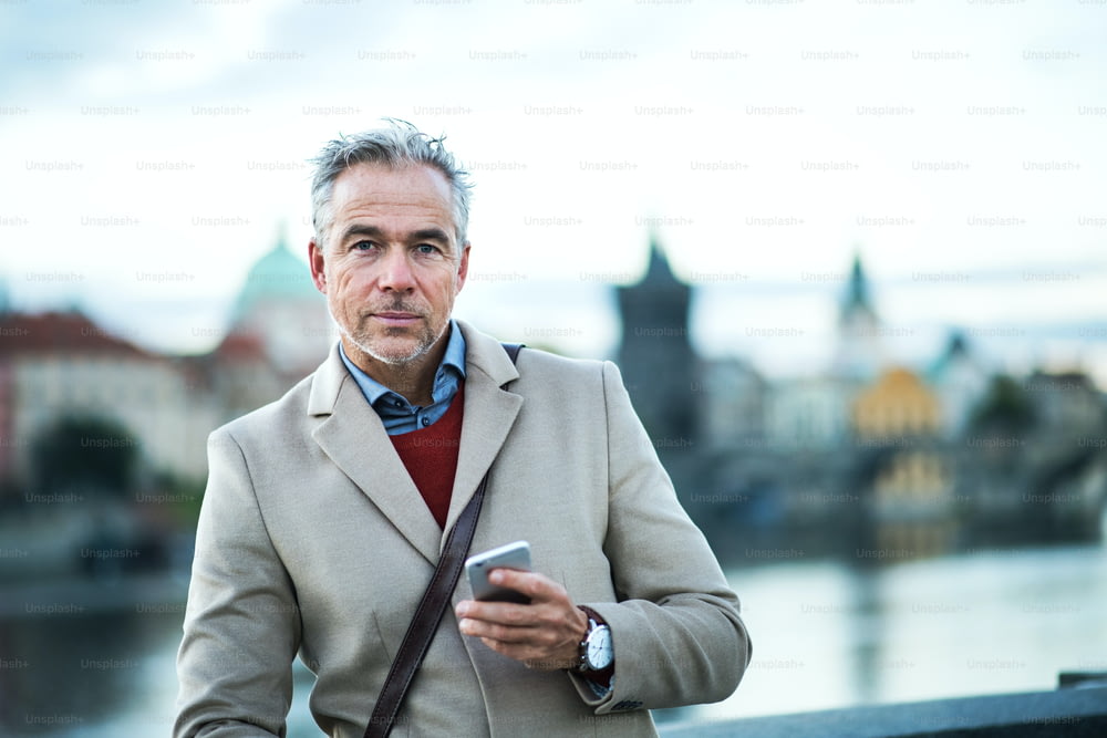 Hombre de negocios guapo maduro con teléfono inteligente de pie junto al río Vltava en la ciudad de Praga. Espacio de copia.