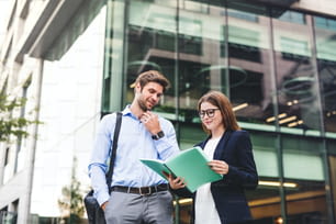 Un giovane uomo d'affari e una donna d'affari in piedi di fronte a un edificio, guardando appunti e parlando.