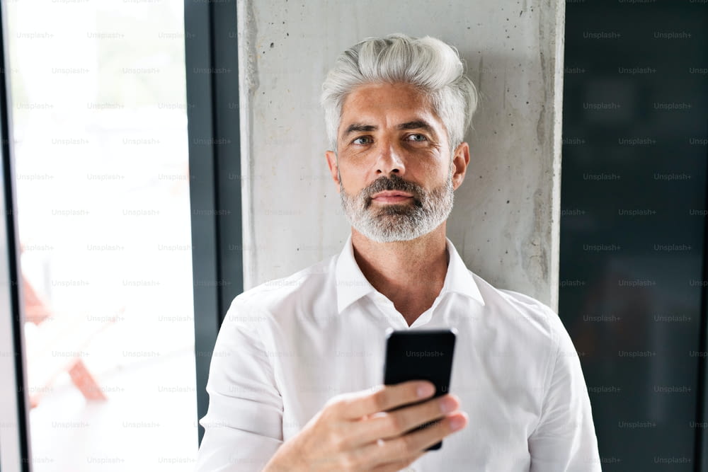 Mature businessman in white shirt in the office standing at the concrete wall, holding a smartphone, texting.