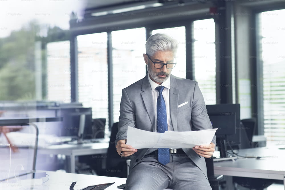 Uomo d'affari maturo in abito grigio seduto alla scrivania in ufficio, leggendo documenti.