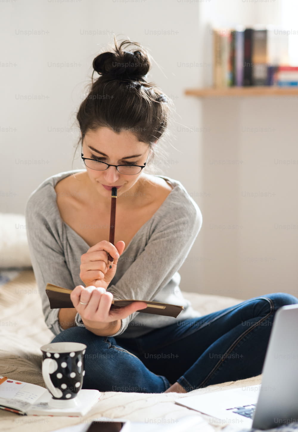 Bela jovem mulher sentada na cama, trabalhando em laptop, escrevendo algo em seu caderno, home office.